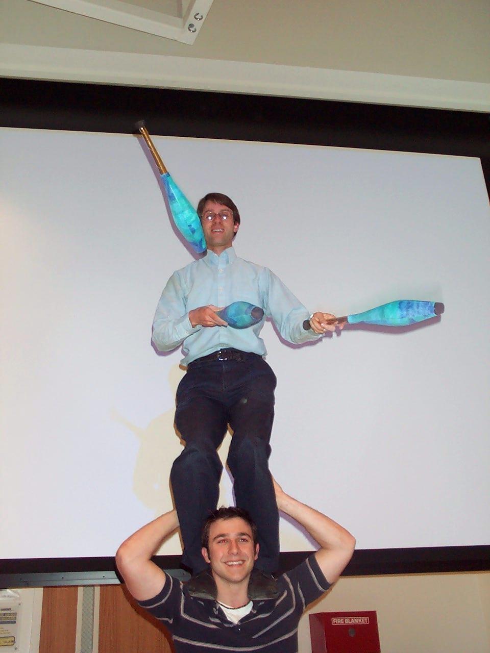 Dr. Deardorff juggling 3 clubs while standing on the shoulders of a student